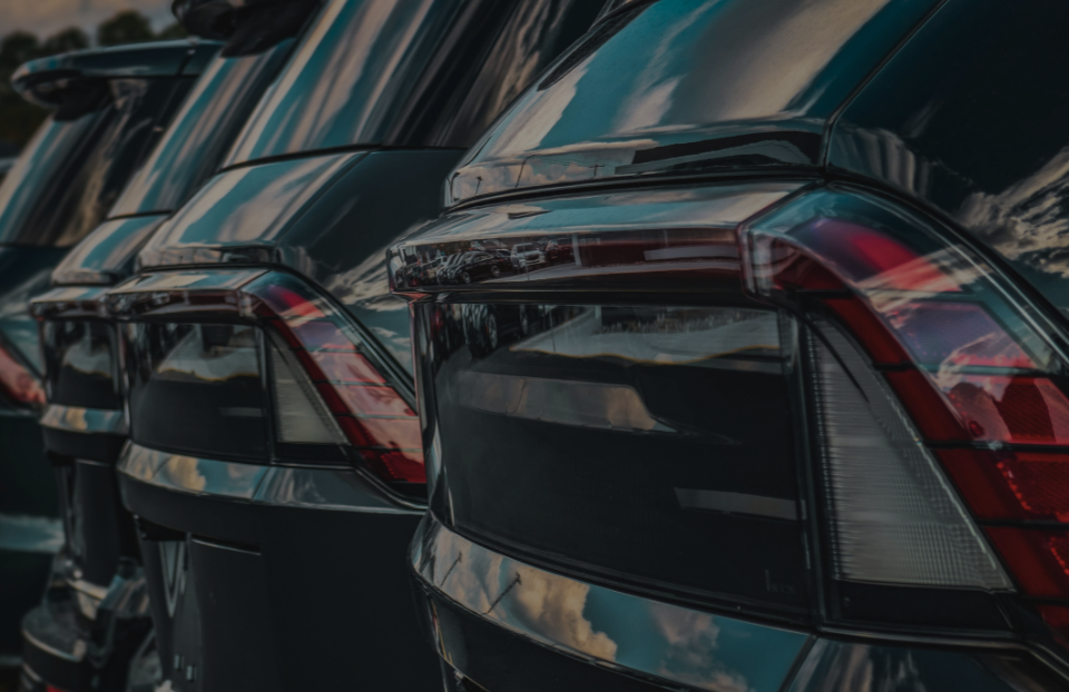 Close-up of the rear ends of dark cars parked in a row, reflecting the sky and surroundings in their glossy surfaces.