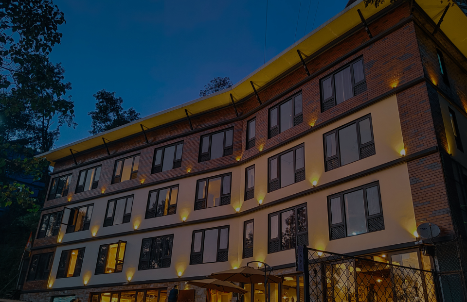A five-story brick hotel-like building with a curved facade, illuminated by warm wall lights at dusk under a deep blue sky, with trees on the left and a gated ground-floor area.